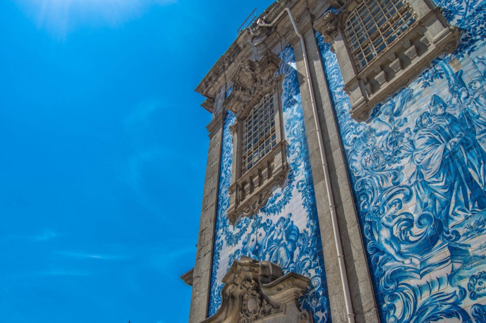 Fotografia de uma igreja no Porto com fachada revestida de azulejos azuis e brancos, capturada sob um céu azul claro. A imagem destaca os detalhes artísticos e arquitetónicos típicos das igrejas portuguesas. Ideal para promover as igrejas e catedrais mais bonitas do Porto, enfatizando a rica herança cultural e religiosa da cidade.