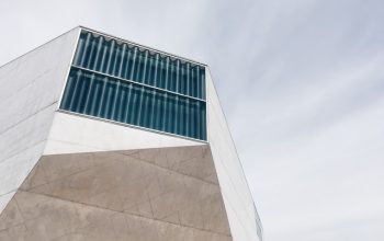 Fotografia da Casa da Música no Porto, um edifício moderno com linhas angulares e grandes janelas de vidro, destacando-se contra um céu parcialmente nublado. A imagem capta a arquitetura icónica e contemporânea deste famoso centro cultural. Ideal para promover a agenda de eventos e atividades na Casa da Música, no Porto